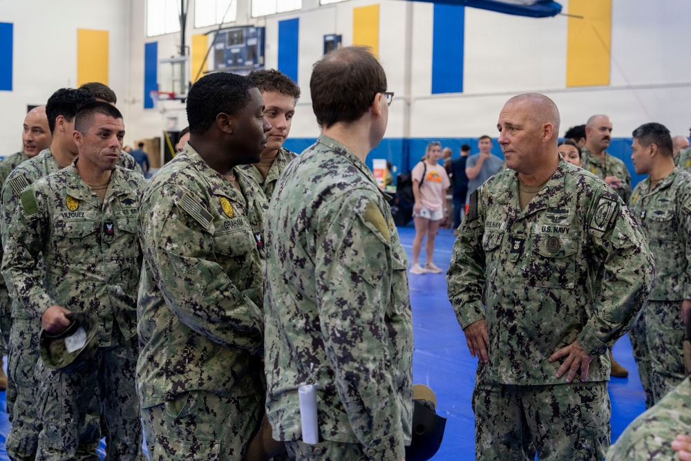 A group of U.S. Navy personnel in NWU Type I (digital camouflage) uniforms are gathered inside what appears to be a gymnasium or indoor sports facility. A senior officer or senior enlisted member on the right, whose name tape reads 'KULBAN' or similar, appears to be engaged in a briefing or conversation with several sailors. The setting suggests a pre-deployment muster, training event, or official visit.