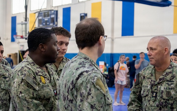 A group of U.S. Navy personnel in NWU Type I (digital camouflage) uniforms are gathered inside what appears to be a gymnasium or indoor sports facility. A senior officer or senior enlisted member on the right, whose name tape reads 'KULBAN' or similar, appears to be engaged in a briefing or conversation with several sailors. The setting suggests a pre-deployment muster, training event, or official visit.
