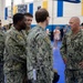 A group of U.S. Navy personnel in NWU Type I (digital camouflage) uniforms are gathered inside what appears to be a gymnasium or indoor sports facility. A senior officer or senior enlisted member on the right, whose name tape reads 'KULBAN' or similar, appears to be engaged in a briefing or conversation with several sailors. The setting suggests a pre-deployment muster, training event, or official visit.