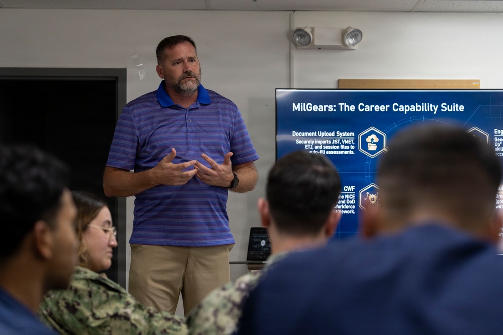 A civilian presenter is delivering a briefing on 'MilGears: The Career Capability Suite' to a group of military personnel in Navy uniforms. The presentation is displayed on a large screen behind him, highlighting features such as a Document Upload System and CWF framework. The setting appears to be a conference or training room on a military installation.