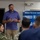 A civilian presenter is delivering a briefing on 'MilGears: The Career Capability Suite' to a group of military personnel in Navy uniforms. The presentation is displayed on a large screen behind him, highlighting features such as a Document Upload System and CWF framework. The setting appears to be a conference or training room on a military installation.