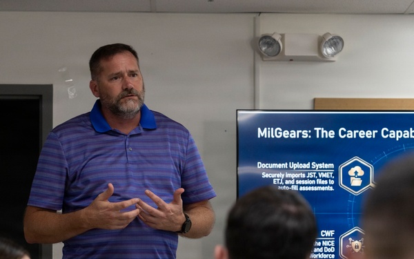 A civilian presenter is delivering a briefing on 'MilGears: The Career Capability Suite' to a group of military personnel in Navy uniforms. The presentation is displayed on a large screen behind him, highlighting features such as a Document Upload System and CWF framework. The setting appears to be a conference or training room on a military installation.