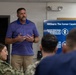 A civilian presenter is delivering a briefing on 'MilGears: The Career Capability Suite' to a group of military personnel in Navy uniforms. The presentation is displayed on a large screen behind him, highlighting features such as a Document Upload System and CWF framework. The setting appears to be a conference or training room on a military installation.