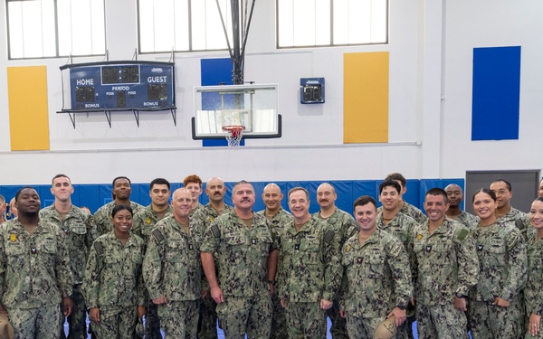 A large group of approximately 25 U.S. Navy personnel in NWU Type III (woodland camouflage) uniforms pose for a formal group photograph inside a military or school gymnasium. The personnel include both men and women of diverse backgrounds, standing in a single row on a blue gymnasium mat. The setting features a basketball court with a scoreboard, blue and gold wall padding, and high windows.