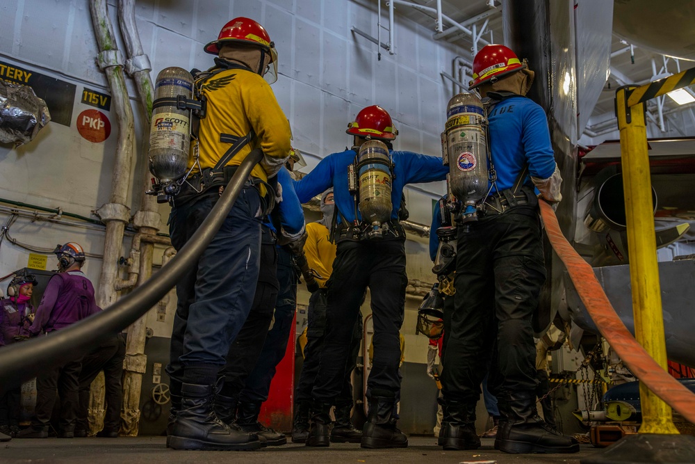 Three personnel wearing red hard hats and Scott Air-Pak SCBA (self-contained breathing apparatus) units are positioned in what appears to be a ship's interior compartment, likely during a damage control or firefighting drill. They are dressed in color-coded shirts (yellow and blue) over dark pants and are holding or standing near large hoses. Additional personnel in purple gear are visible in the background.