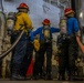 Three personnel wearing red hard hats and Scott Air-Pak SCBA (self-contained breathing apparatus) units are positioned in what appears to be a ship's interior compartment, likely during a damage control or firefighting drill. They are dressed in color-coded shirts (yellow and blue) over dark pants and are holding or standing near large hoses. Additional personnel in purple gear are visible in the background.