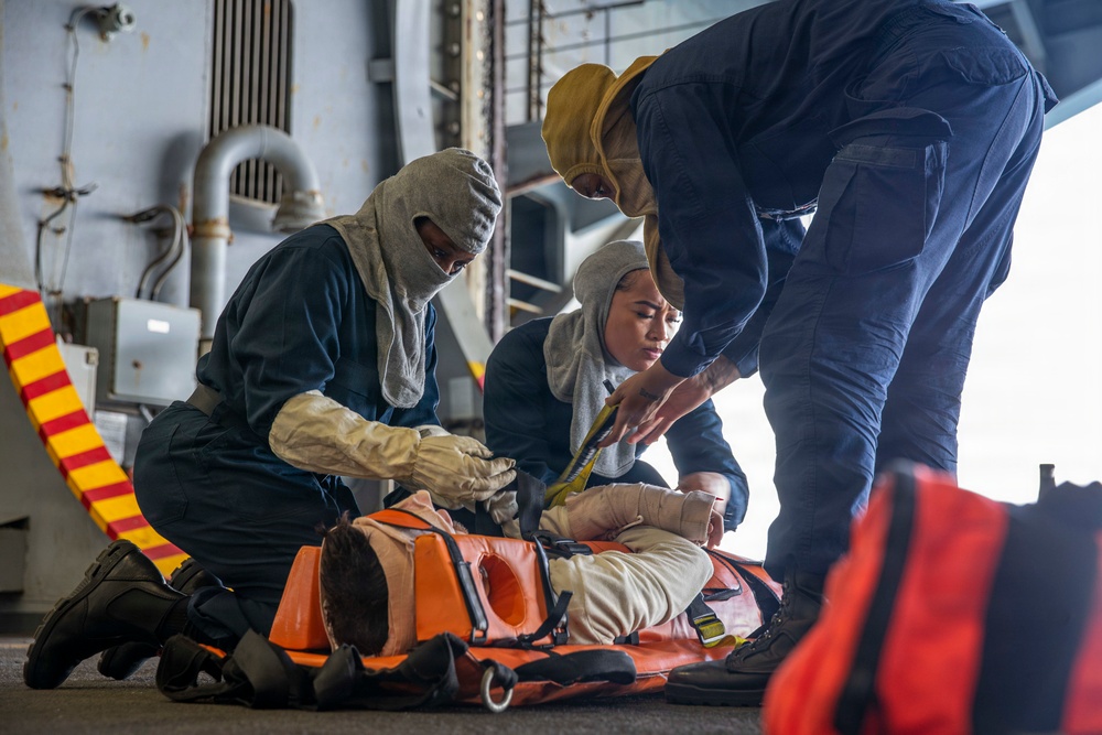 Three personnel in navy blue coveralls are conducting what appears to be a medical or casualty evacuation drill aboard a naval vessel. They are securing a person onto an orange Stokes litter (rescue stretcher) using straps. Two of the personnel are wearing flash hoods and one wears a yellow hard hat, indicating this is a damage control or emergency response training exercise.