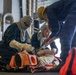 Three personnel in navy blue coveralls are conducting what appears to be a medical or casualty evacuation drill aboard a naval vessel. They are securing a person onto an orange Stokes litter (rescue stretcher) using straps. Two of the personnel are wearing flash hoods and one wears a yellow hard hat, indicating this is a damage control or emergency response training exercise.