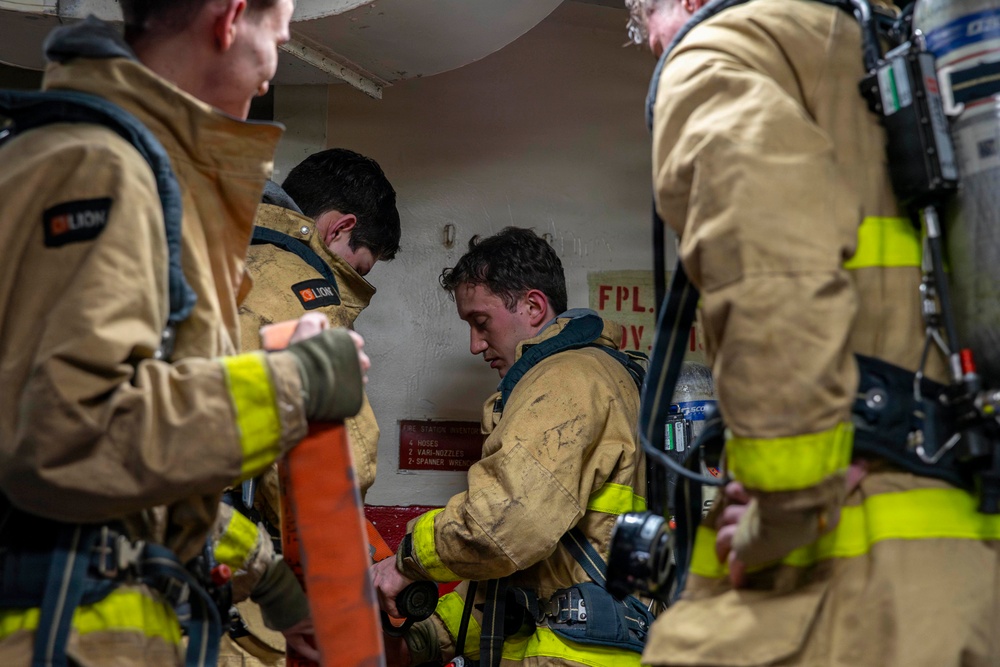 A group of firefighters in turnout gear are gathered in a confined space, appearing to be inside a vessel or industrial facility. They are wearing tan and yellow reflective protective gear branded 'LION' and are equipped with SCBA (self-contained breathing apparatus) units. The scene appears to be a training exercise or operational briefing near a fire station equipment panel.