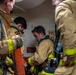 A group of firefighters in turnout gear are gathered in a confined space, appearing to be inside a vessel or industrial facility. They are wearing tan and yellow reflective protective gear branded 'LION' and are equipped with SCBA (self-contained breathing apparatus) units. The scene appears to be a training exercise or operational briefing near a fire station equipment panel.