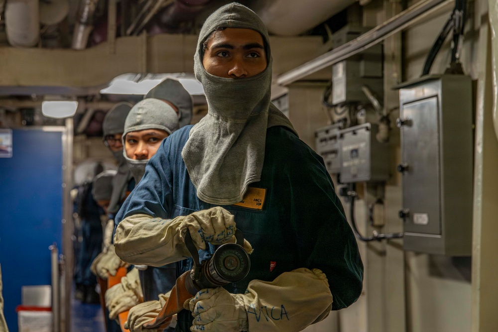 Two individuals wearing gray fire-resistant balaclavas and protective gloves are positioned in what appears to be a naval vessel interior corridor. The person in the foreground is holding a fire hose nozzle, suggesting participation in damage control or firefighting drill training. The setting, protective gear, and equipment are consistent with U.S. Navy shipboard emergency response procedures.