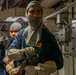 Two individuals wearing gray fire-resistant balaclavas and protective gloves are positioned in what appears to be a naval vessel interior corridor. The person in the foreground is holding a fire hose nozzle, suggesting participation in damage control or firefighting drill training. The setting, protective gear, and equipment are consistent with U.S. Navy shipboard emergency response procedures.