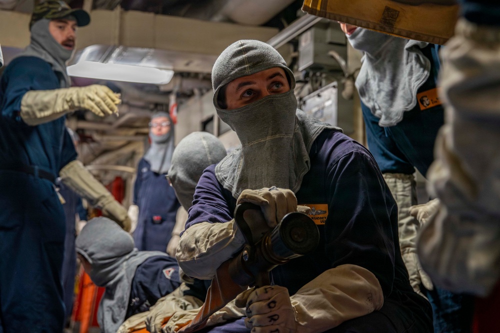 A group of personnel in navy blue coveralls and protective gear, including flash hoods (balaclavas) and heat-resistant gloves, are engaged in what appears to be a damage control or firefighting drill aboard a naval vessel. The central figure is holding a fire hose nozzle and wearing a name tag, with multiple crew members visible in the confined interior space. The setting appears to be below decks on a military ship.