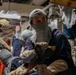 A group of personnel in navy blue coveralls and protective gear, including flash hoods (balaclavas) and heat-resistant gloves, are engaged in what appears to be a damage control or firefighting drill aboard a naval vessel. The central figure is holding a fire hose nozzle and wearing a name tag, with multiple crew members visible in the confined interior space. The setting appears to be below decks on a military ship.