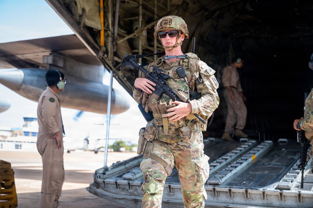A fully equipped military service member in Operational Camouflage Pattern (OCP) uniform stands at the rear cargo ramp of a large military transport aircraft, likely a C-130 or similar. The soldier is armed with what appears to be an M4 carbine and is wearing full tactical gear including a helmet, plate carrier, and sunglasses. Additional personnel in various uniforms are visible in the background near the aircraft.
