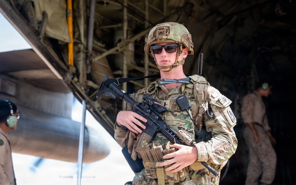 A fully equipped military service member in Operational Camouflage Pattern (OCP) uniform stands at the rear cargo ramp of a large military transport aircraft, likely a C-130 or similar. The soldier is armed with what appears to be an M4 carbine and is wearing full tactical gear including a helmet, plate carrier, and sunglasses. Additional personnel in various uniforms are visible in the background near the aircraft.