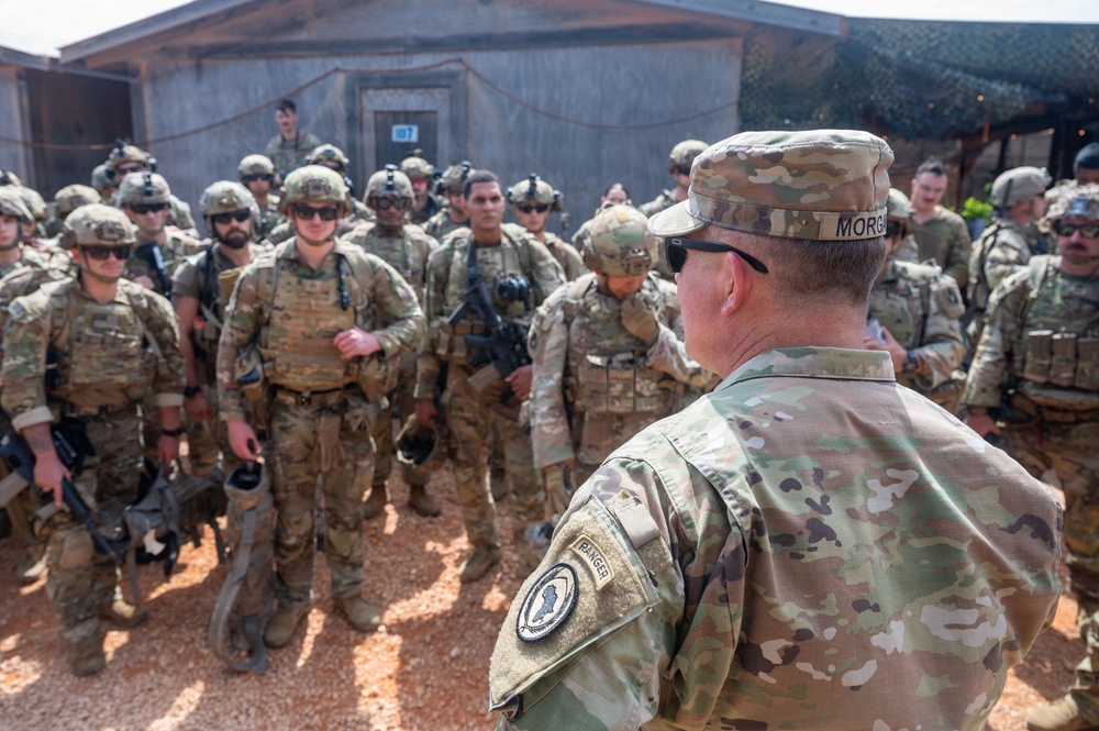 A senior U.S. Army officer with the last name 'Morgan,' wearing a Ranger tab and a unit patch, addresses a large formation of soldiers in OCP (Operational Camouflage Pattern) uniforms at what appears to be a forward operating base or training facility. The soldiers are fully equipped with body armor, helmets, and weapons. The setting appears to be an arid or semi-arid environment based on the red-dirt ground and the building structures visible in the background.