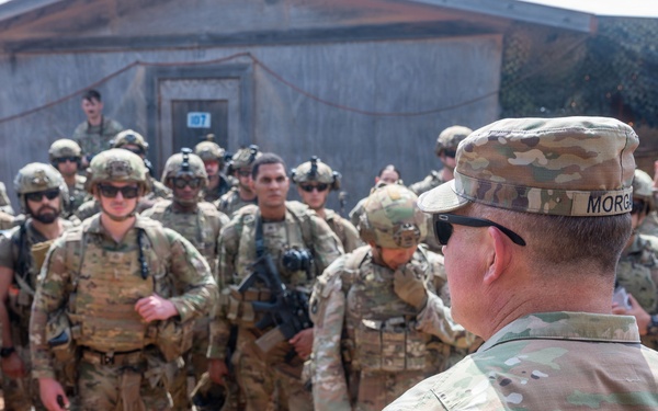 A senior U.S. Army officer with the last name 'Morgan,' wearing a Ranger tab and a unit patch, addresses a large formation of soldiers in OCP (Operational Camouflage Pattern) uniforms at what appears to be a forward operating base or training facility. The soldiers are fully equipped with body armor, helmets, and weapons. The setting appears to be an arid or semi-arid environment based on the red-dirt ground and the building structures visible in the background.