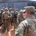 A senior U.S. Army officer with the last name 'Morgan,' wearing a Ranger tab and a unit patch, addresses a large formation of soldiers in OCP (Operational Camouflage Pattern) uniforms at what appears to be a forward operating base or training facility. The soldiers are fully equipped with body armor, helmets, and weapons. The setting appears to be an arid or semi-arid environment based on the red-dirt ground and the building structures visible in the background.