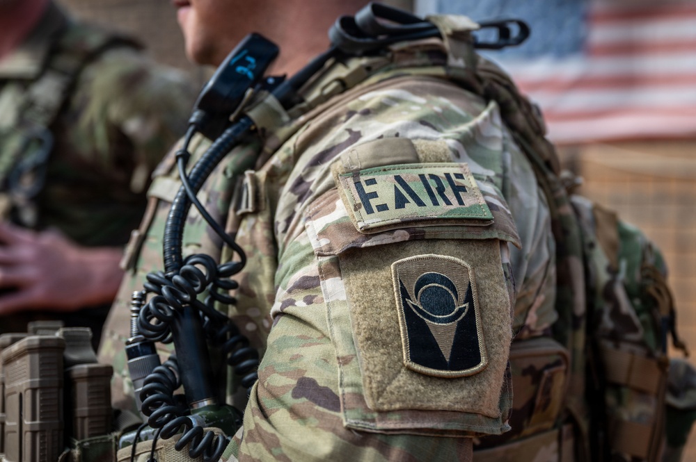 Close-up photograph of a U.S. military service member's left shoulder area showing tactical gear and unit patches. The soldier is wearing MultiCam (OCP) camouflage uniform with a plate carrier/chest rig and communications equipment. Two distinct patches are visible on the sleeve, along with a blurred American flag in the background.