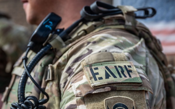 Close-up photograph of a U.S. military service member's left shoulder area showing tactical gear and unit patches. The soldier is wearing MultiCam (OCP) camouflage uniform with a plate carrier/chest rig and communications equipment. Two distinct patches are visible on the sleeve, along with a blurred American flag in the background.