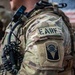 Close-up photograph of a U.S. military service member's left shoulder area showing tactical gear and unit patches. The soldier is wearing MultiCam (OCP) camouflage uniform with a plate carrier/chest rig and communications equipment. Two distinct patches are visible on the sleeve, along with a blurred American flag in the background.