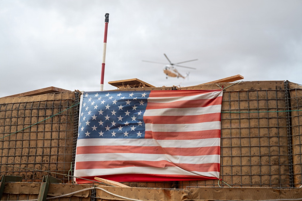 An American flag is displayed on a wire mesh barrier wall, consistent with military forward operating base (FOB) construction using HESCO-style barriers filled with earth. A helicopter is visible in the overcast sky in the background. The setting strongly suggests a military installation or combat outpost.