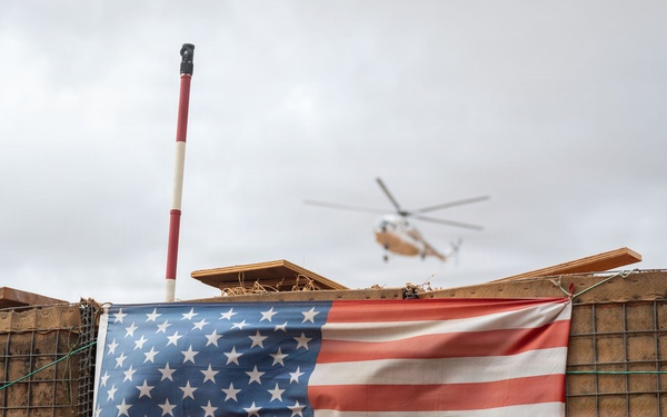 An American flag is displayed on a wire mesh barrier wall, consistent with military forward operating base (FOB) construction using HESCO-style barriers filled with earth. A helicopter is visible in the overcast sky in the background. The setting strongly suggests a military installation or combat outpost.