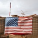 An American flag is displayed on a wire mesh barrier wall, consistent with military forward operating base (FOB) construction using HESCO-style barriers filled with earth. A helicopter is visible in the overcast sky in the background. The setting strongly suggests a military installation or combat outpost.
