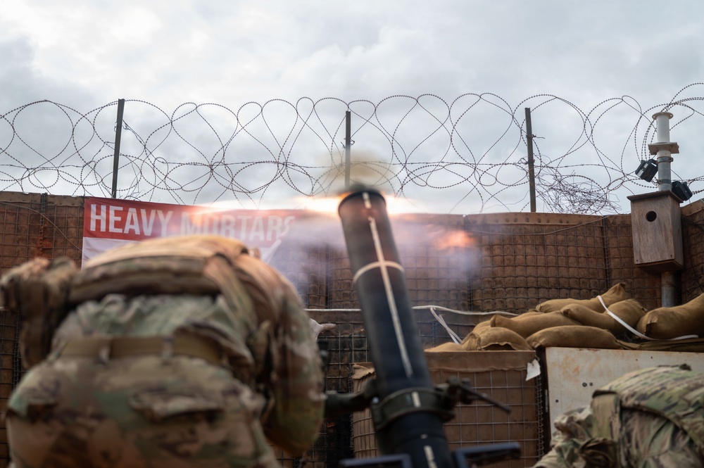 A military mortar is being fired from a fortified position, with visible muzzle blast and flame exiting the tube. A soldier in camouflage uniform and tactical gear is visible in the foreground. The position is surrounded by sandbags, a HESCO barrier wall, and concertina/razor wire, with a red banner reading 'HEAVY MORTARS' visible in the background.