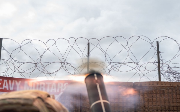 A military mortar is being fired from a fortified position, with visible muzzle blast and flame exiting the tube. A soldier in camouflage uniform and tactical gear is visible in the foreground. The position is surrounded by sandbags, a HESCO barrier wall, and concertina/razor wire, with a red banner reading 'HEAVY MORTARS' visible in the background.