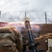 A military mortar is being fired from a fortified position, with visible muzzle blast and flame exiting the tube. A soldier in camouflage uniform and tactical gear is visible in the foreground. The position is surrounded by sandbags, a HESCO barrier wall, and concertina/razor wire, with a red banner reading 'HEAVY MORTARS' visible in the background.