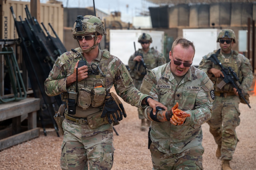The image shows U.S. Army soldiers in a military base or training environment. A soldier identified by the name tape 'BLACKBURN' appears to have simulated or actual injuries to his hands, while a fellow soldier holds a radio and assists him. Additional armed soldiers are visible in the background.