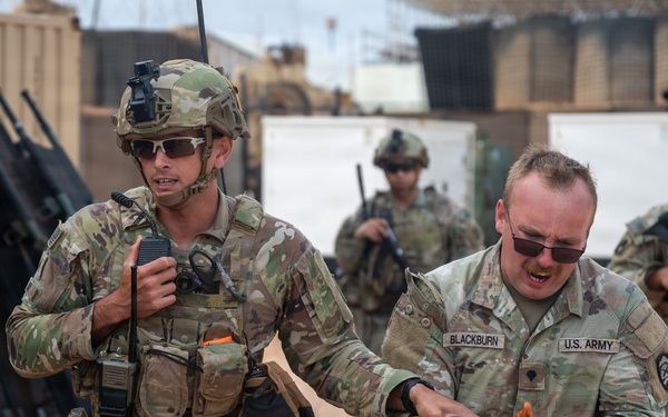 The image shows U.S. Army soldiers in a military base or training environment. A soldier identified by the name tape 'BLACKBURN' appears to have simulated or actual injuries to his hands, while a fellow soldier holds a radio and assists him. Additional armed soldiers are visible in the background.