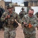 The image shows U.S. Army soldiers in a military base or training environment. A soldier identified by the name tape 'BLACKBURN' appears to have simulated or actual injuries to his hands, while a fellow soldier holds a radio and assists him. Additional armed soldiers are visible in the background.