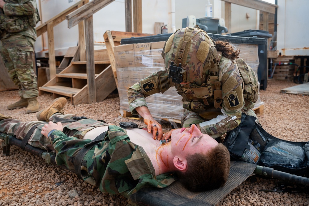 A military medic in OCP (Operational Camouflage Pattern) uniform and full tactical gear is performing a medical treatment or assessment on a simulated casualty lying on a litter/stretcher on a gravel surface. The casualty, wearing woodland camouflage BDUs with an exposed chest, has simulated wound makeup (orange/red moulage) applied to the neck and torso area. This appears to be a combat casualty care training exercise based on the controlled environment and simulated injuries.