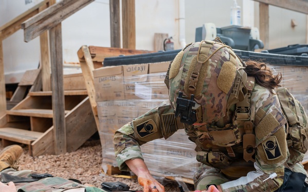 A military medic in OCP (Operational Camouflage Pattern) uniform and full tactical gear is performing a medical treatment or assessment on a simulated casualty lying on a litter/stretcher on a gravel surface. The casualty, wearing woodland camouflage BDUs with an exposed chest, has simulated wound makeup (orange/red moulage) applied to the neck and torso area. This appears to be a combat casualty care training exercise based on the controlled environment and simulated injuries.
