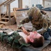 A military medic in OCP (Operational Camouflage Pattern) uniform and full tactical gear is performing a medical treatment or assessment on a simulated casualty lying on a litter/stretcher on a gravel surface. The casualty, wearing woodland camouflage BDUs with an exposed chest, has simulated wound makeup (orange/red moulage) applied to the neck and torso area. This appears to be a combat casualty care training exercise based on the controlled environment and simulated injuries.