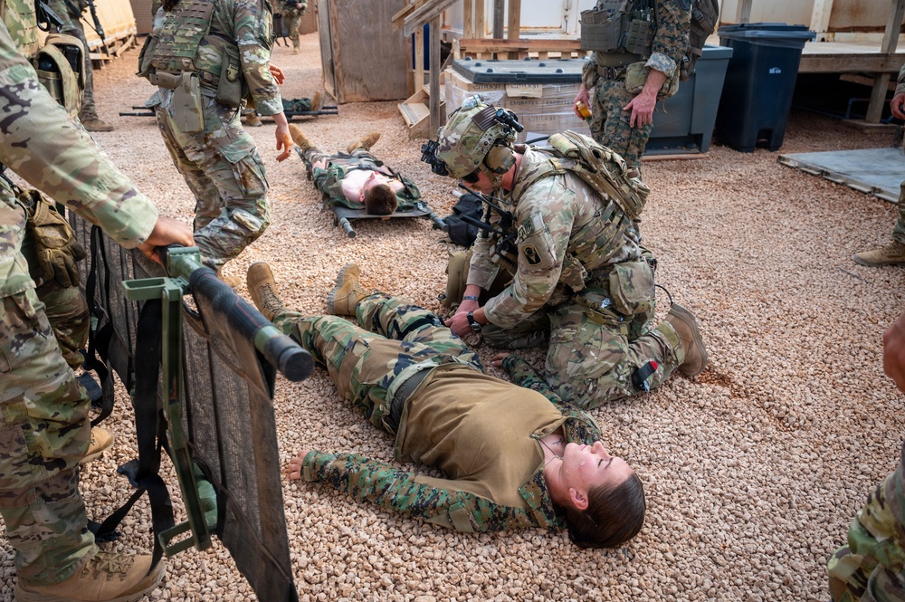 Military personnel are conducting a combat casualty care or medical evacuation training exercise. Soldiers in OCP (Operational Camouflage Pattern) uniforms are treating simulated casualties wearing MARPAT (Marine Pattern) uniforms lying on a gravel surface. A litter/stretcher is visible, and multiple 'casualties' are positioned on the ground in what appears to be a controlled training environment.