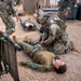 Military personnel are conducting a combat casualty care or medical evacuation training exercise. Soldiers in OCP (Operational Camouflage Pattern) uniforms are treating simulated casualties wearing MARPAT (Marine Pattern) uniforms lying on a gravel surface. A litter/stretcher is visible, and multiple 'casualties' are positioned on the ground in what appears to be a controlled training environment.
