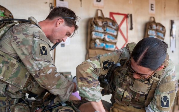 Two military personnel in OCP (Operational Camouflage Pattern) uniforms are performing tactical combat casualty care (TCCC) on a simulated or actual casualty lying on a litter/stretcher. Both individuals are wearing full tactical gear including plate carriers, and one is wearing blue medical gloves while treating the patient's lower extremities. The setting appears to be an indoor training or aid station environment with medical supplies visible on the wall in the background.