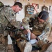 Two military personnel in OCP (Operational Camouflage Pattern) uniforms are performing tactical combat casualty care (TCCC) on a simulated or actual casualty lying on a litter/stretcher. Both individuals are wearing full tactical gear including plate carriers, and one is wearing blue medical gloves while treating the patient's lower extremities. The setting appears to be an indoor training or aid station environment with medical supplies visible on the wall in the background.