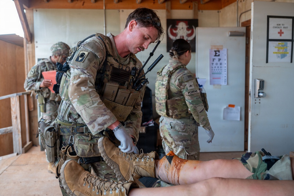 Military medical personnel are conducting a tactical combat casualty care (TCCC) training exercise in what appears to be a field aid station. A soldier wearing full combat gear and white medical gloves is treating a simulated leg wound on a casualty, while other uniformed personnel are present in the background. Medical reference materials, including an eye chart and a 'How to Contact FAST' poster with a Red Cross symbol, are visible on the wall.