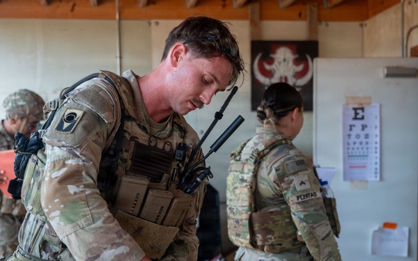 Military medical personnel are conducting a tactical combat casualty care (TCCC) training exercise in what appears to be a field aid station. A soldier wearing full combat gear and white medical gloves is treating a simulated leg wound on a casualty, while other uniformed personnel are present in the background. Medical reference materials, including an eye chart and a 'How to Contact FAST' poster with a Red Cross symbol, are visible on the wall.