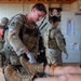 Military medical personnel are conducting a tactical combat casualty care (TCCC) training exercise in what appears to be a field aid station. A soldier wearing full combat gear and white medical gloves is treating a simulated leg wound on a casualty, while other uniformed personnel are present in the background. Medical reference materials, including an eye chart and a 'How to Contact FAST' poster with a Red Cross symbol, are visible on the wall.