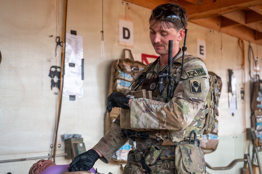A U.S. Army combat medic in MultiCam/OCP uniform is attending to a patient (partially visible at bottom) in what appears to be a field medical treatment station or aid station. The soldier is wearing black medical gloves, has a stethoscope around his neck, carries a radio, and appears to be checking his watch while treating the patient. The setting includes labeled treatment bays (D and E visible) and medical supplies mounted on the walls.