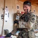 A U.S. Army combat medic in MultiCam/OCP uniform is attending to a patient (partially visible at bottom) in what appears to be a field medical treatment station or aid station. The soldier is wearing black medical gloves, has a stethoscope around his neck, carries a radio, and appears to be checking his watch while treating the patient. The setting includes labeled treatment bays (D and E visible) and medical supplies mounted on the walls.