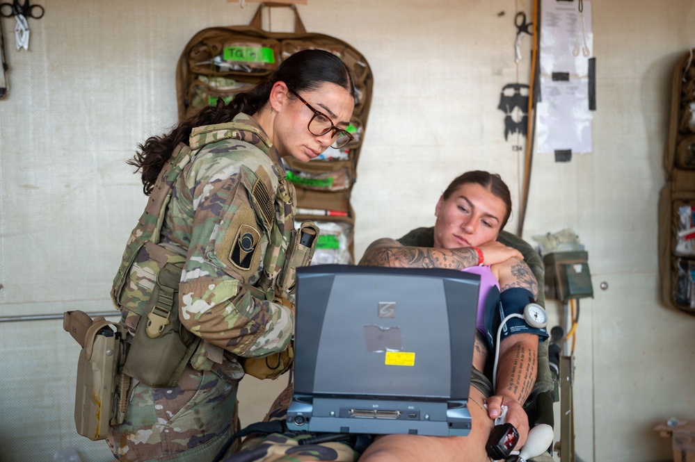 A military medic in full combat gear and glasses reviews data on a ruggedized laptop while attending to a patient seated in a medical treatment area. The patient, wearing a blood pressure cuff on their arm, appears relaxed and has visible tattoos. The setting appears to be a field medical station or aid station with medical supplies mounted on the wall.