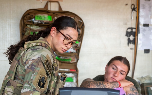 A military medic in full combat gear and glasses reviews data on a ruggedized laptop while attending to a patient seated in a medical treatment area. The patient, wearing a blood pressure cuff on their arm, appears relaxed and has visible tattoos. The setting appears to be a field medical station or aid station with medical supplies mounted on the wall.