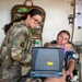 A military medic in full combat gear and glasses reviews data on a ruggedized laptop while attending to a patient seated in a medical treatment area. The patient, wearing a blood pressure cuff on their arm, appears relaxed and has visible tattoos. The setting appears to be a field medical station or aid station with medical supplies mounted on the wall.